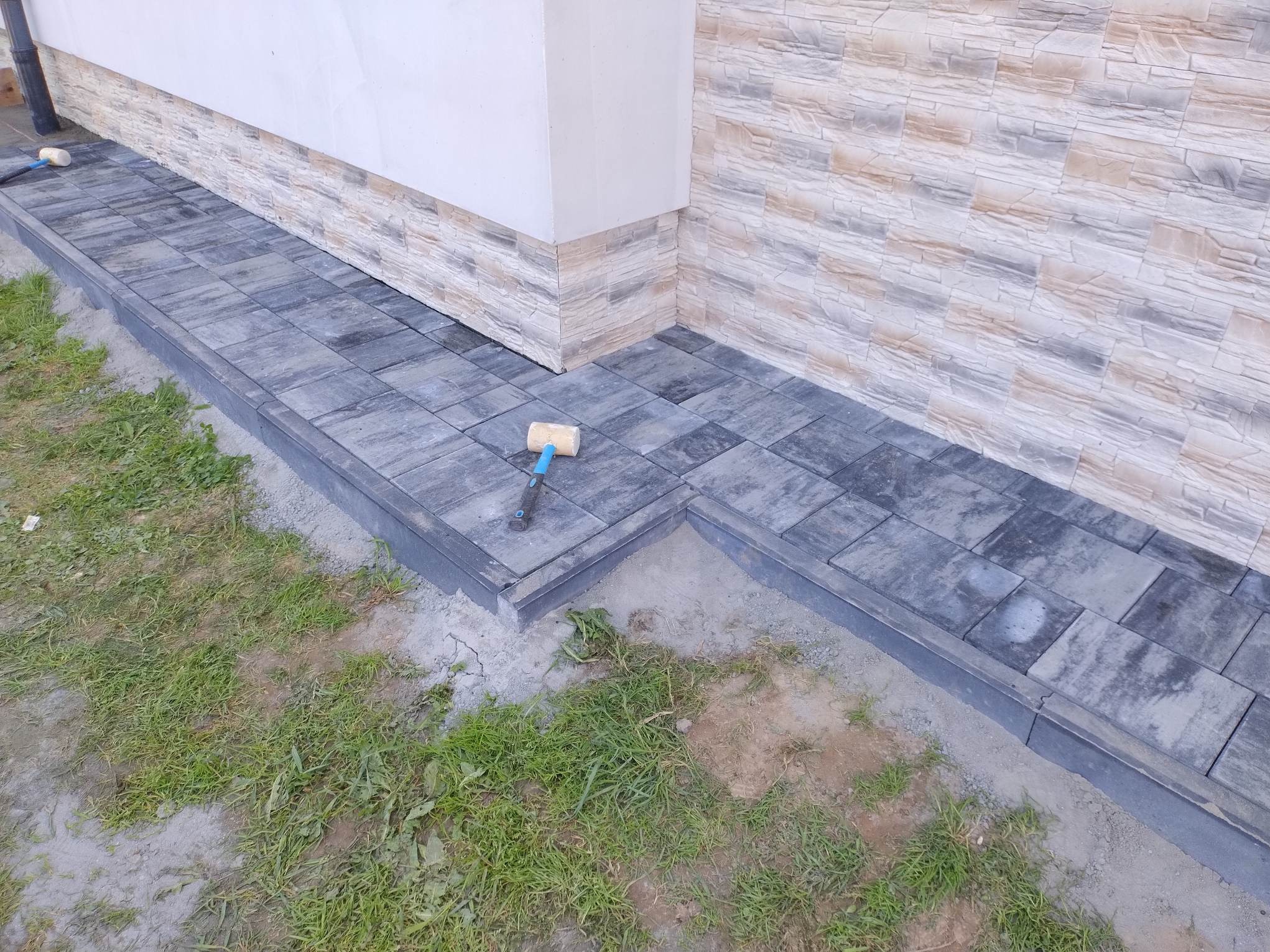 Ułożona, szara kostka brukowa przy elewacji budynku z dekoracyjnym kamieniem. Gumowy młotek leży na kostce. Widoczne trawnik i obrzeża.