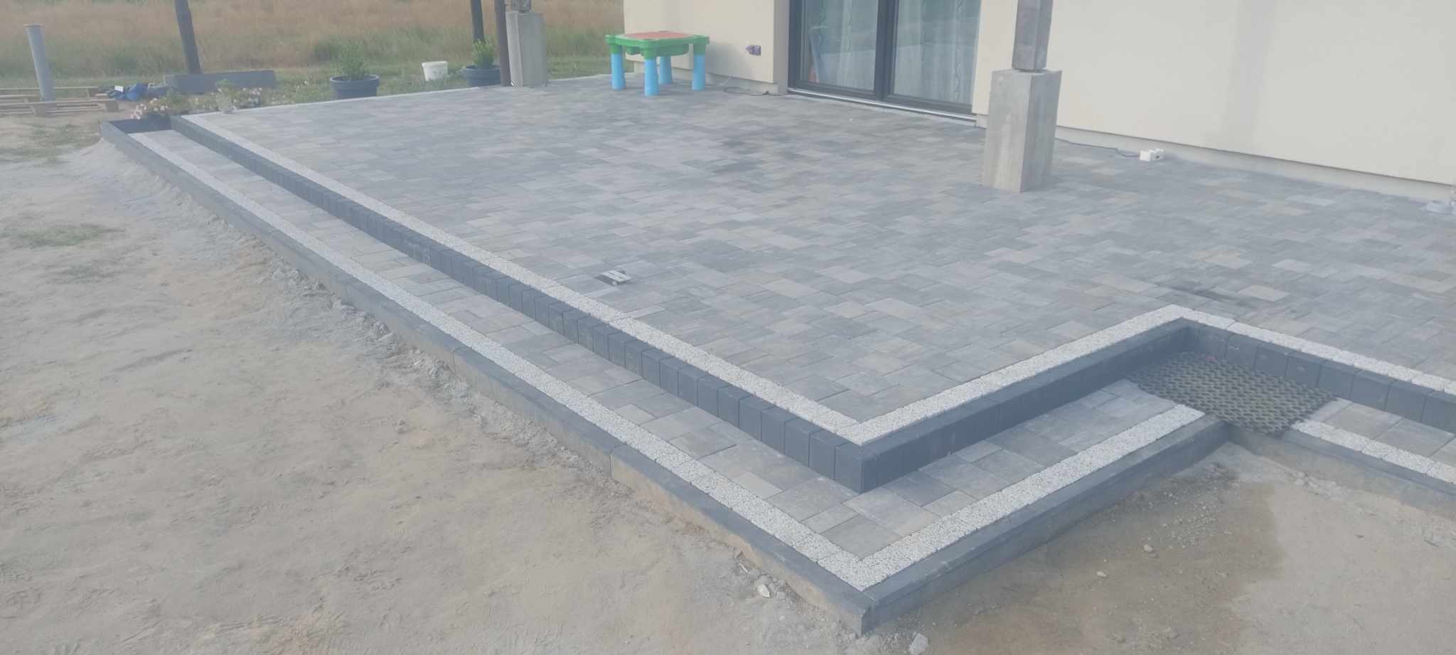 Nowoczesny taras z szarej kostki brukowej, wykończony obrzeżami z jasnego grysu. Schody z kostki prowadzą na taras. W tle dom z przesuwnymi drzwiami.