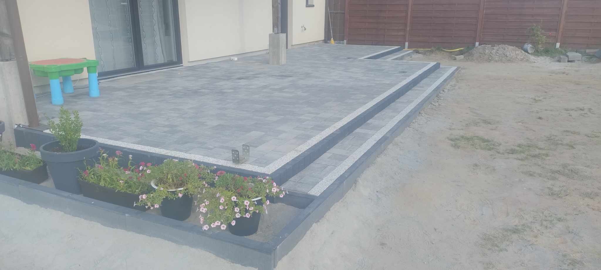 Szara kostka brukowa ułożona na tarasie z dwoma stopniami, obrzeża z jasnego grysu, donice z kwiatami, drewniany płot w tle. Widok z poziomu gruntu.