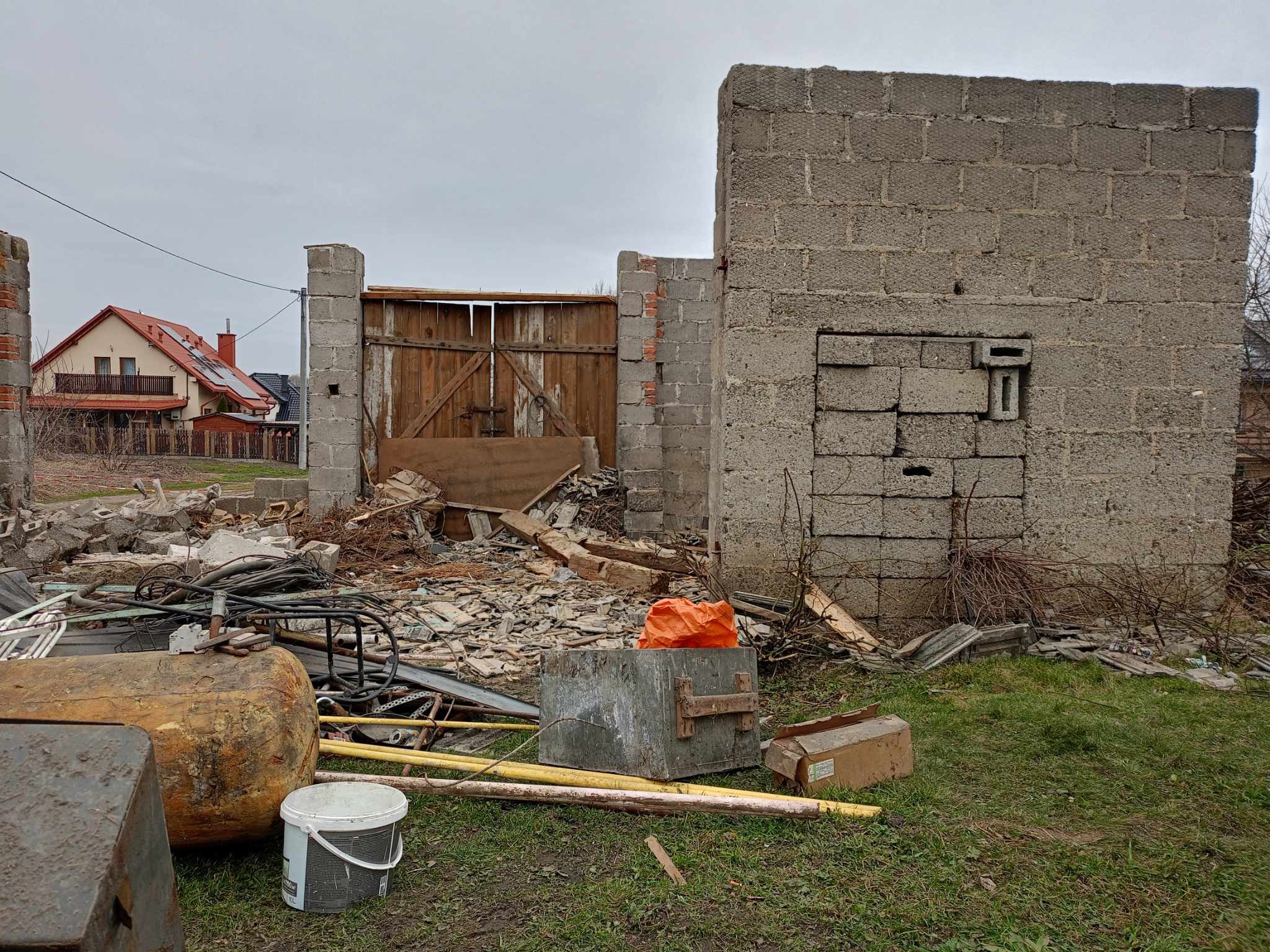 Po rozbiórce: fragment muru z pustaków, drewniana brama i gruzowisko na trawie. W tle dom z czerwoną dachówką pod pochmurnym niebem.
