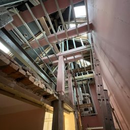 SEG - Skośny kadr na konstrukcję sufitu podwieszanego z metalowych profili i drewnianych belek, widoczne fragmenty różowej ściany i drabina oparta o ścianę.