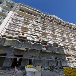 SegmentPRO - Budynek w trakcie ocieplania, osłonięty rusztowaniami. Widoczni pracownicy w pomarańczowych kombinezonach. Na dole materiały budowlane i narzędzia.