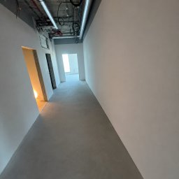 SegmentPRO - Długi korytarz w trakcie wykończenia z gładkimi białymi ścianami, szarą posadzką i widocznymi instalacjami wentylacyjnymi oraz elektrycznymi pod sufitem.
