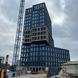 SegmentPRO - Nowoczesny budynek biurowy w trakcie budowy, z ciemną elewacją i przeszklonymi piętrami, obok wysokiego żurawia budowlanego. Część konstrukcji jest w surowym betonie, a na pierwszym planie widać...