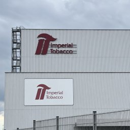 SegmentPRO - Budynek przemysłowy z falistej blachy z dwoma logotypami Imperial Tobacco, większym bezpośrednio na elewacji i mniejszym na białym panelu. Widoczne metalowe schody ewakuacyjne i ogrodzenie z paletami.