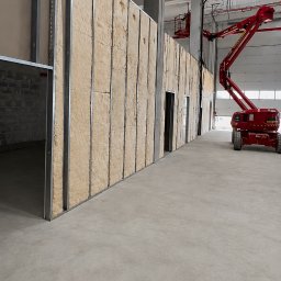 SegmentPRO - Montaż konstrukcji ściany działowej z metalowych profili wypełnionych wełną mineralną w przestronnej hali, z widocznym podnośnikiem koszowym w tle.
