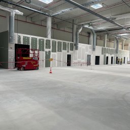 SegmentPRO - Duża hala w trakcie prac wykończeniowych: ściany z zielonych płyt gipsowo-kartonowych z częściowym szpachlowaniem, widoczne kanały wentylacyjne i podnośnik nożycowy.