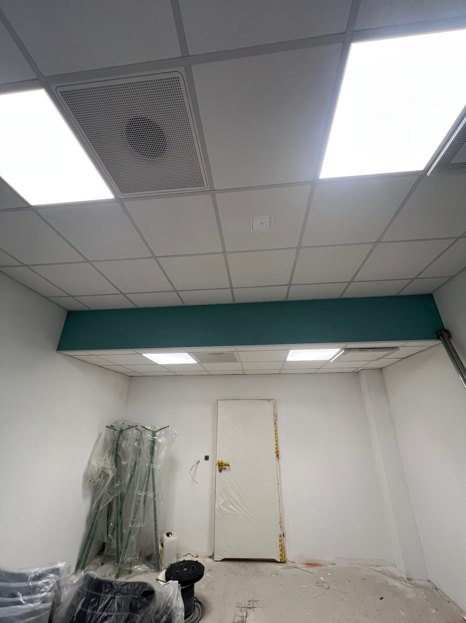 Biały sufit kasetonowy z oświetleniem LED i kratką wentylacyjną w nowym biurze. Ściany białe, widoczna zielona listwa i drzwi zabezpieczone folią. W tle sprzęt budowlany.