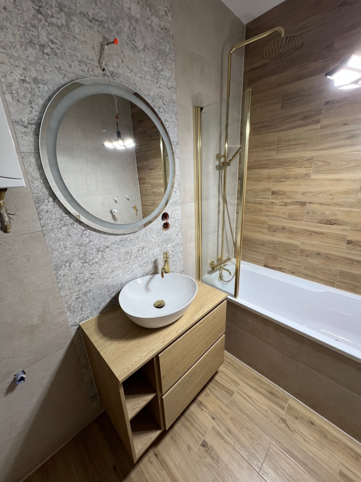 Wykończona łazienka z okrągłym lustrem LED, umywalką nablatową na drewnianej szafce i wanną z parawanem. Płytki imitujące drewno na podłodze i ścianach.