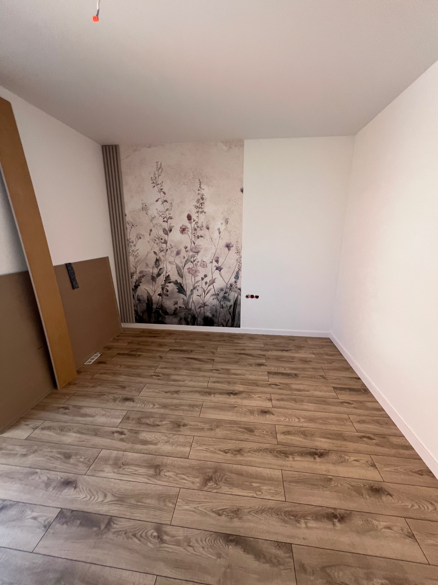 Pokój po remoncie z panelami podłogowymi imitującymi drewno, ozdobną tapetą w kwiaty i listwami dekoracyjnymi. Częściowo widoczne płyty gipsowe.