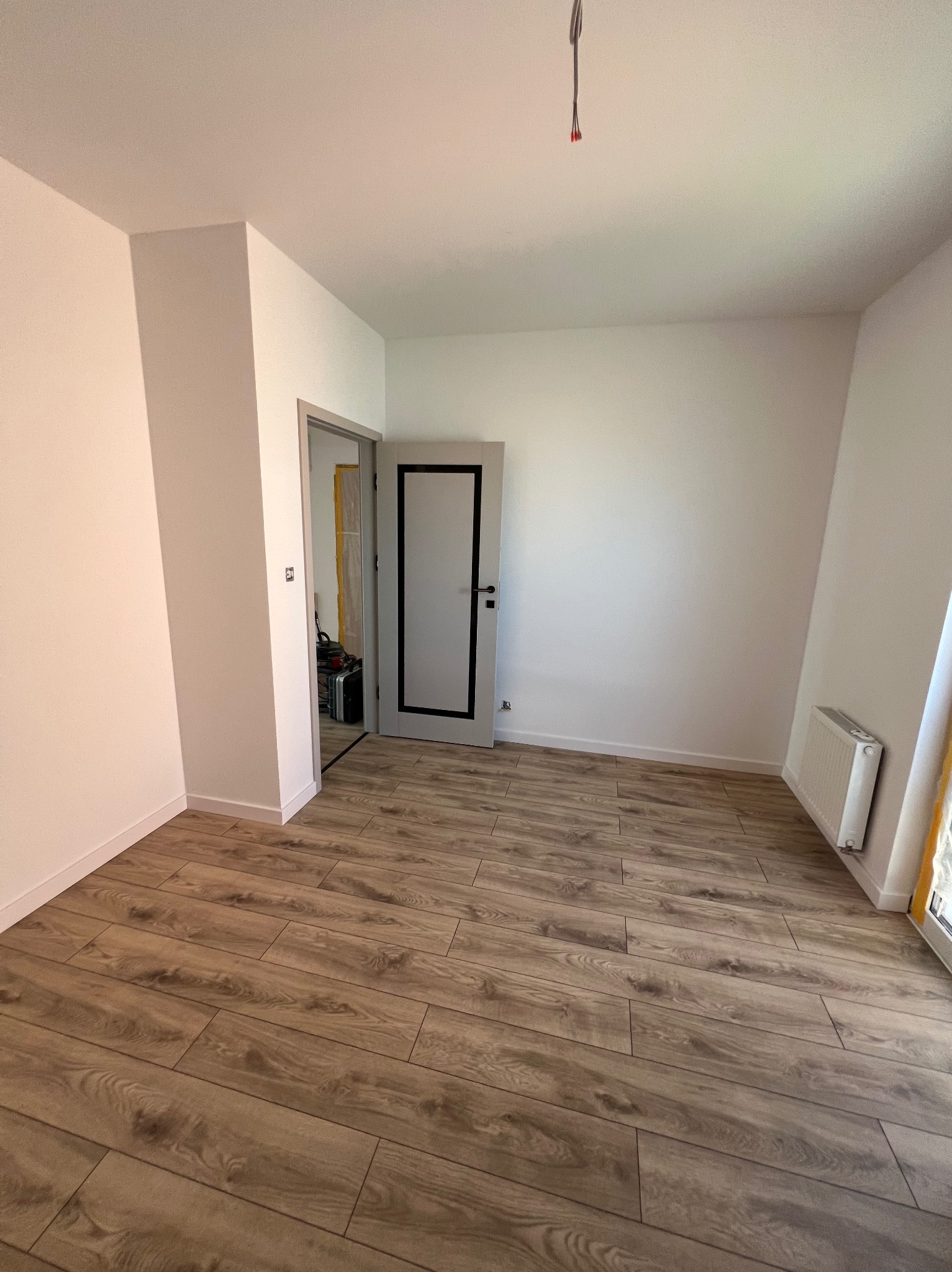 Jasne wnętrze po remoncie: drewnopodobna podłoga, białe ściany, grzejnik i drzwi z czarną ramą. Widoczne kable wystające z sufitu.