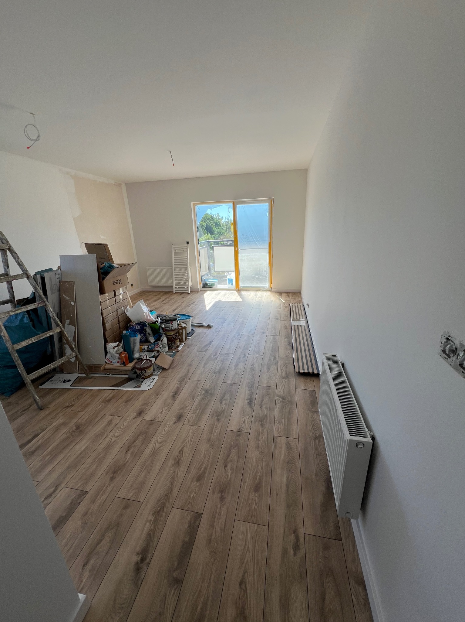 Jasne wnętrze w trakcie remontu: świeżo pomalowane ściany, drewnopodobna podłoga, grzejnik, okno balkonowe i narzędzia malarskie. Widoczne ślady prac wykończeniowych.