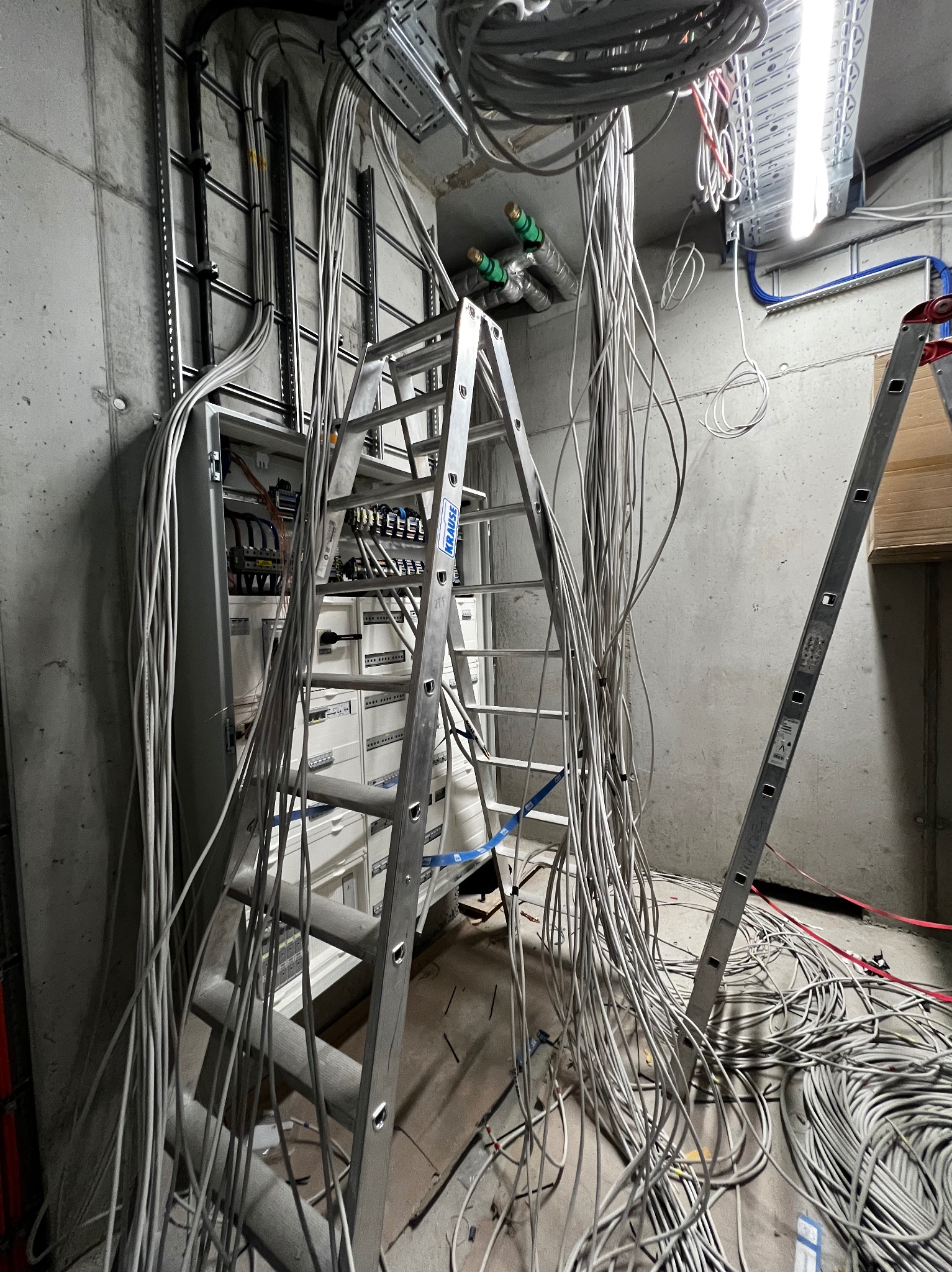 Rozdzielnia elektryczna w surowym wnętrzu, z licznymi kablami i drabiną Krause. Widoczne elementy instalacji elektrycznej na ścianie. Surowy beton w tle.