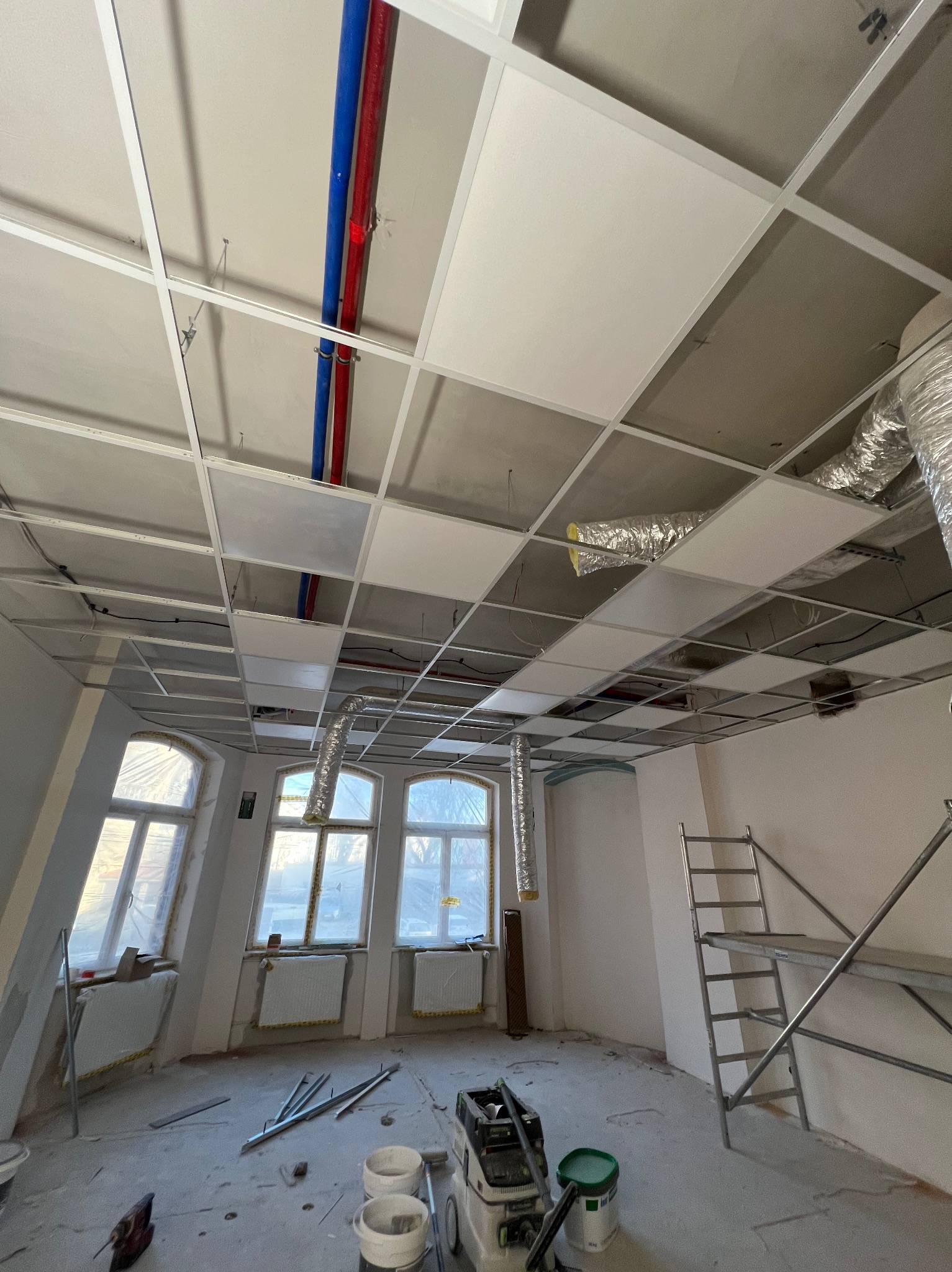 Montaż sufitu podwieszanego kasetonowego w pomieszczeniu z trzema oknami, widoczne rury instalacyjne i elementy wentylacji. Na podłodze narzędzia i drabina.