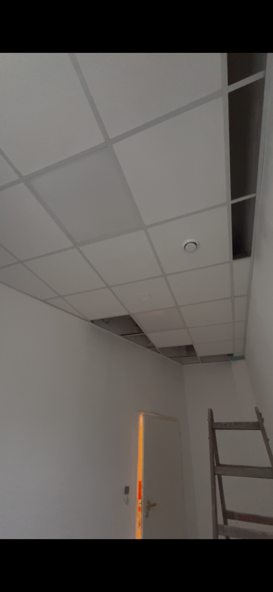 Biały sufit kasetonowy z widocznymi otworami i elementami konstrukcyjnymi, drabina oparta o ścianę, widok z dołu, oświetlenie pomieszczenia.