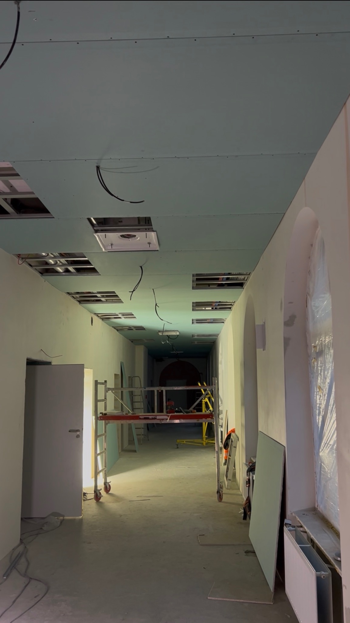 Korytarz w trakcie montażu sufitu z płyt karton-gips, widoczne rusztowania i instalacje. Ściany z łukowymi wnękami, okna zabezpieczone folią.