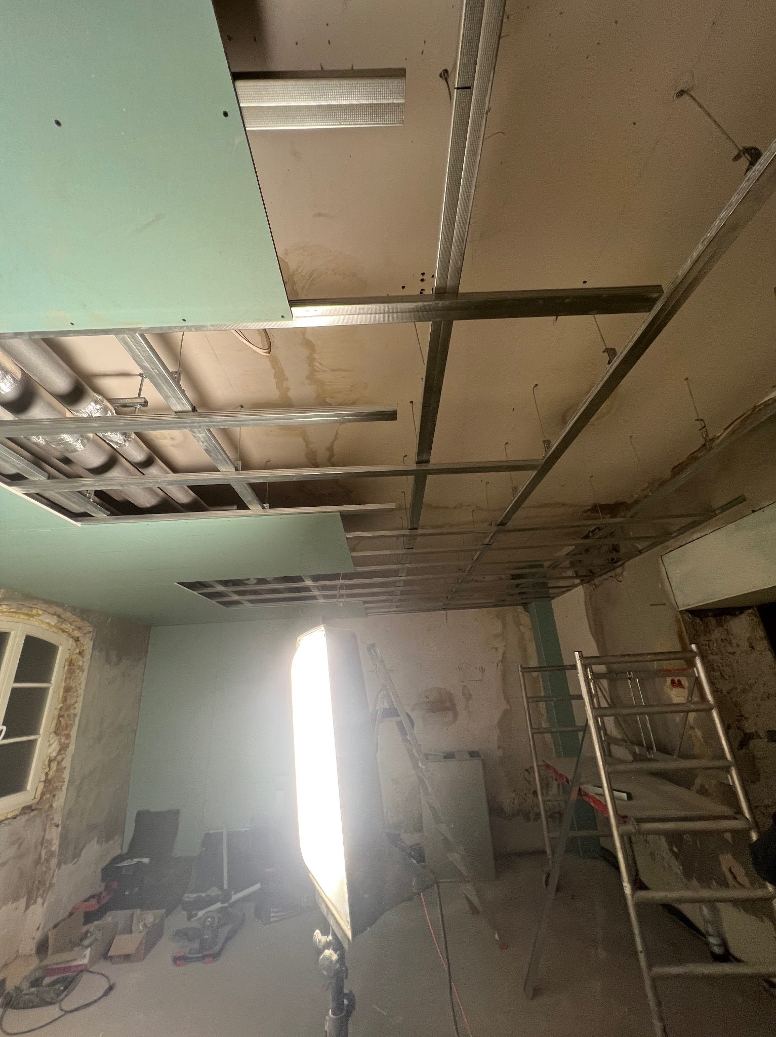 Montaż sufitu podwieszanego z płyt karton-gips na metalowej konstrukcji w remontowanym pomieszczeniu. Widoczne drabiny i narzędzia.