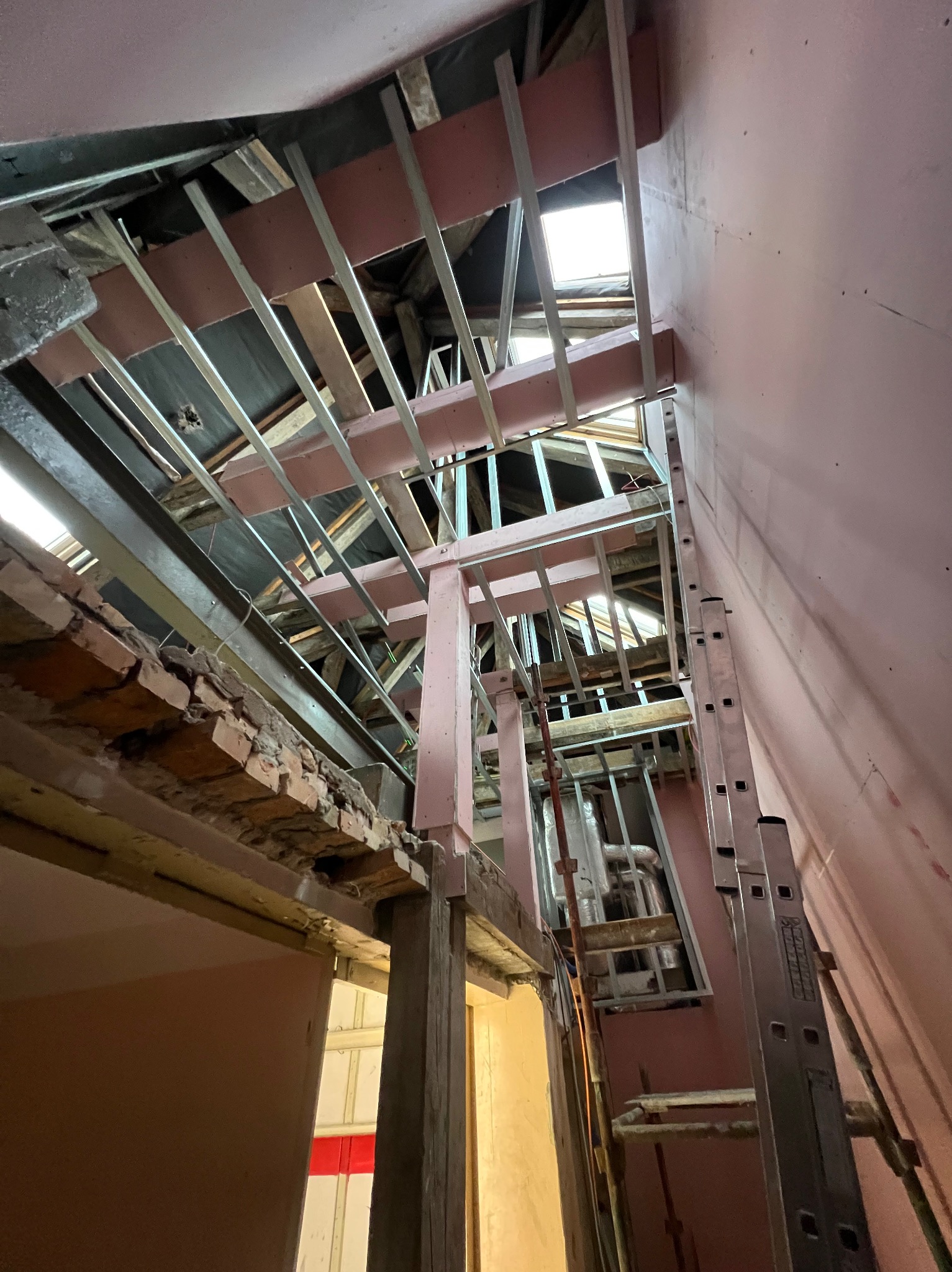 Skośny kadr na konstrukcję sufitu podwieszanego z metalowych profili i drewnianych belek, widoczne fragmenty różowej ściany i drabina oparta o ścianę.