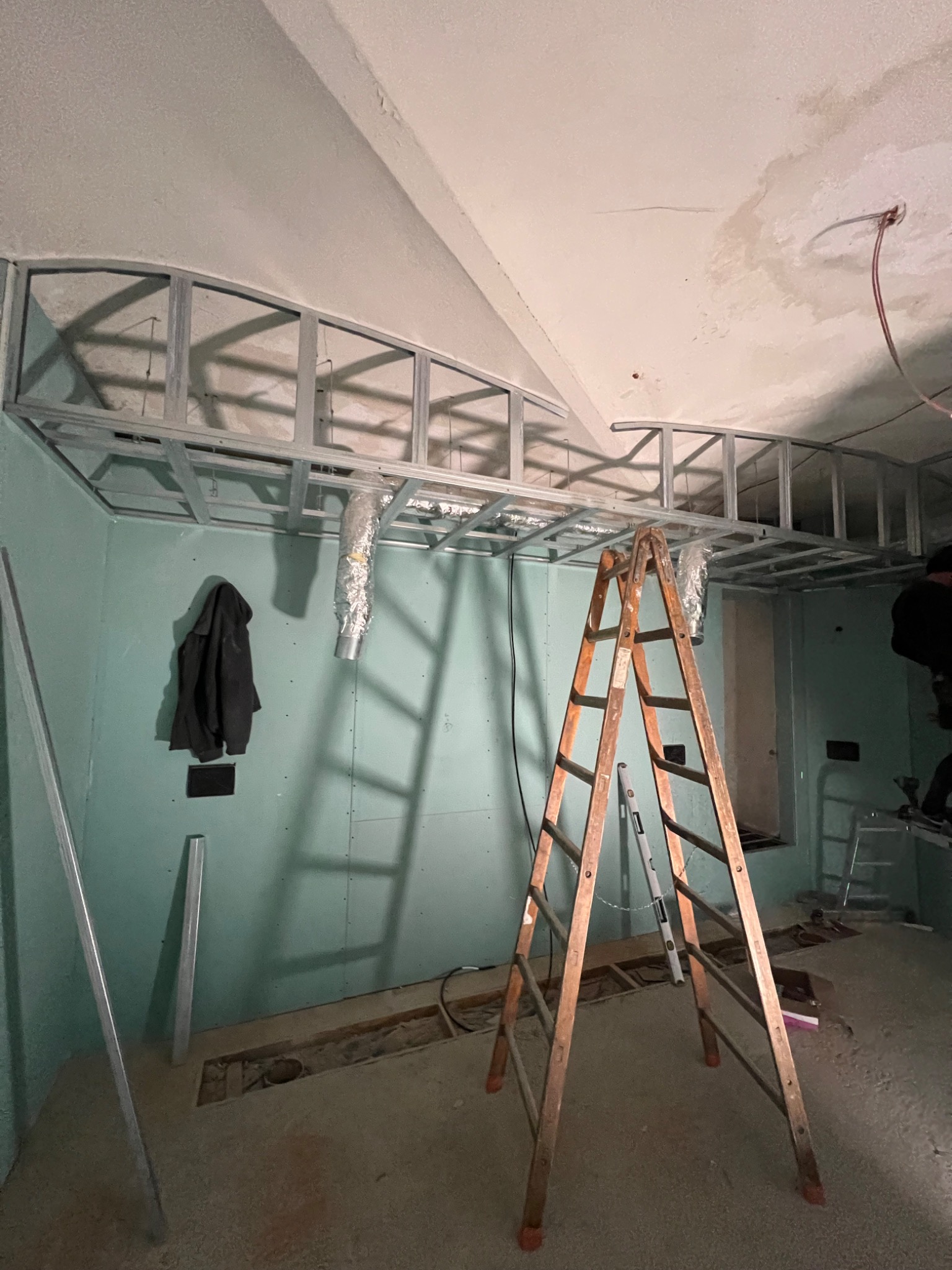 Montaż metalowej konstrukcji sufitu podwieszanego z płyt karton-gips w pomieszczeniu z zielonymi ścianami. Widoczna drabina i elementy wentylacji.