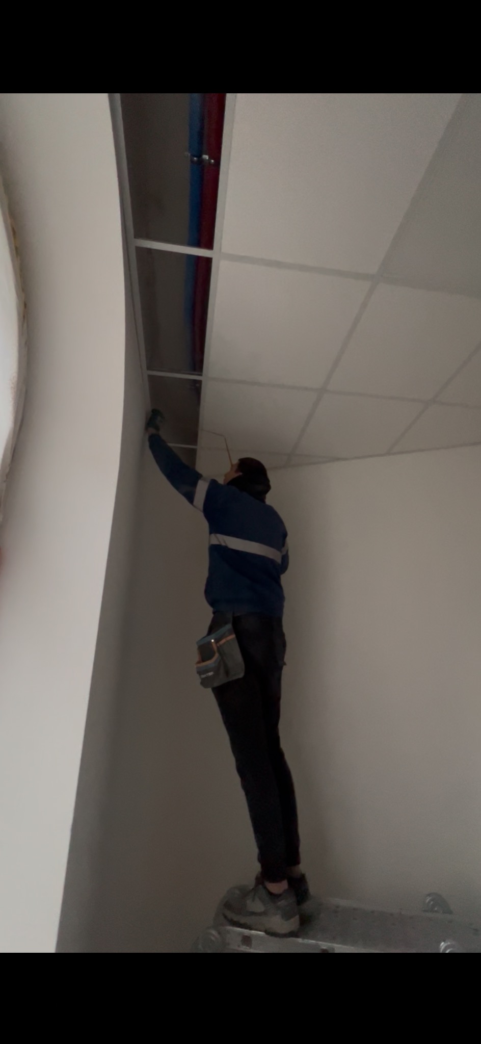 Pracownik na drabinie montuje podwieszany sufit kasetonowy w pomieszczeniu z instalacjami. Widoczne narzędzia i elementy konstrukcyjne sufitu.