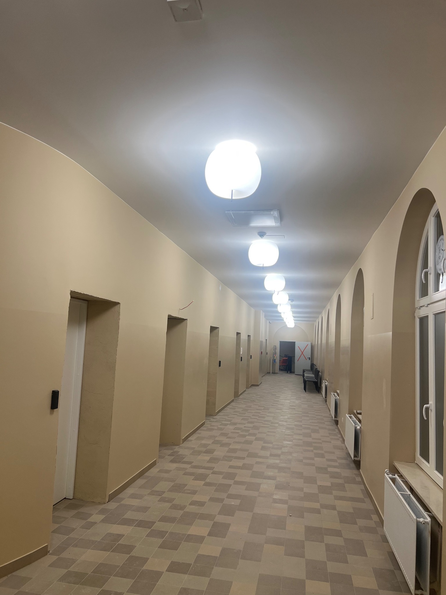 Długi korytarz w budynku po remoncie. Ściany w odcieniach beżu, wzorzysta podłoga, białe drzwi i okna. Sufit z rzędem okrągłych lamp. Widoczne grzejniki.