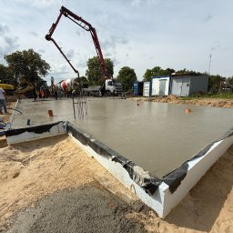 SOVEX SPÓŁKA Z OGRANICZONĄ ODPOWIEDZIALNOŚCIĄ - Wylany fundament pod halę z widoczną pompą do betonu, szalunkiem z izolacją i wystającymi prętami zbrojeniowymi. W tle betoniarka i ekipa budowlana.