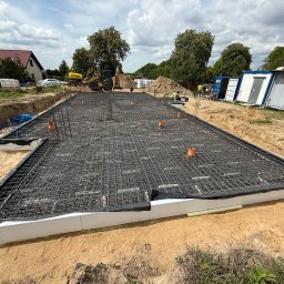 SOVEX SPÓŁKA Z OGRANICZONĄ ODPOWIEDZIALNOŚCIĄ - Zbrojenie fundamentu hali z widocznymi rurami instalacyjnymi, przygotowane do zalania betonem. W tle koparki i kontenery budowlane na placu budowy.