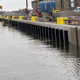 SOVEX SPÓŁKA Z OGRANICZONĄ ODPOWIEDZIALNOŚCIĄ - Nabrzeże portowe w trakcie budowy z żółtymi odbojami i pracownikami w kamizelkach, widok z poziomu wody. W tle widać sprzęt budowlany i elementy infrastruktury portowej.