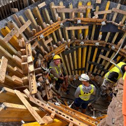 SOVEX SPÓŁKA Z OGRANICZONĄ ODPOWIEDZIALNOŚCIĄ - Budowa fundamentów: pracownicy w kasku i kamizelkach odblaskowych w wykopie z drewnianym szalunkiem, widok z góry na plac budowy.
