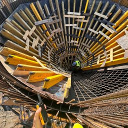 SOVEX SPÓŁKA Z OGRANICZONĄ ODPOWIEDZIALNOŚCIĄ - Wnętrze szalunku zbrojeniowego o nietypowym kształcie, widok z dołu. Pracownik w odblaskowej kamizelce wewnątrz konstrukcji. W tle siatka zbrojeniowa i ręka z radiotelefonem.