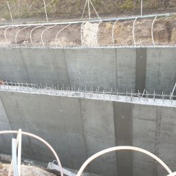 SOVEX SPÓŁKA Z OGRANICZONĄ ODPOWIEDZIALNOŚCIĄ - Betonowa konstrukcja z wystającymi prętami zbrojeniowymi, widok z góry, fragment rury na pierwszym planie, teren budowy w tle.