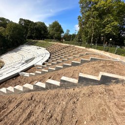 SOVEX SPÓŁKA Z OGRANICZONĄ ODPOWIEDZIALNOŚCIĄ - Budowa amfiteatru: betonowe elementy konstrukcyjne stopniowo wznoszą się na zboczu, otoczone zielenią drzew i błękitem nieba. Widoczny etap prac ziemnych i montażu.