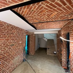 SOVEX SPÓŁKA Z OGRANICZONĄ ODPOWIEDZIALNOŚCIĄ - Ceglany sufit łukowy z czarnymi belkami i białymi panelami. Widoczne ceglane ściany i przejście do pomieszczenia ze schodami. W tle widać schody.