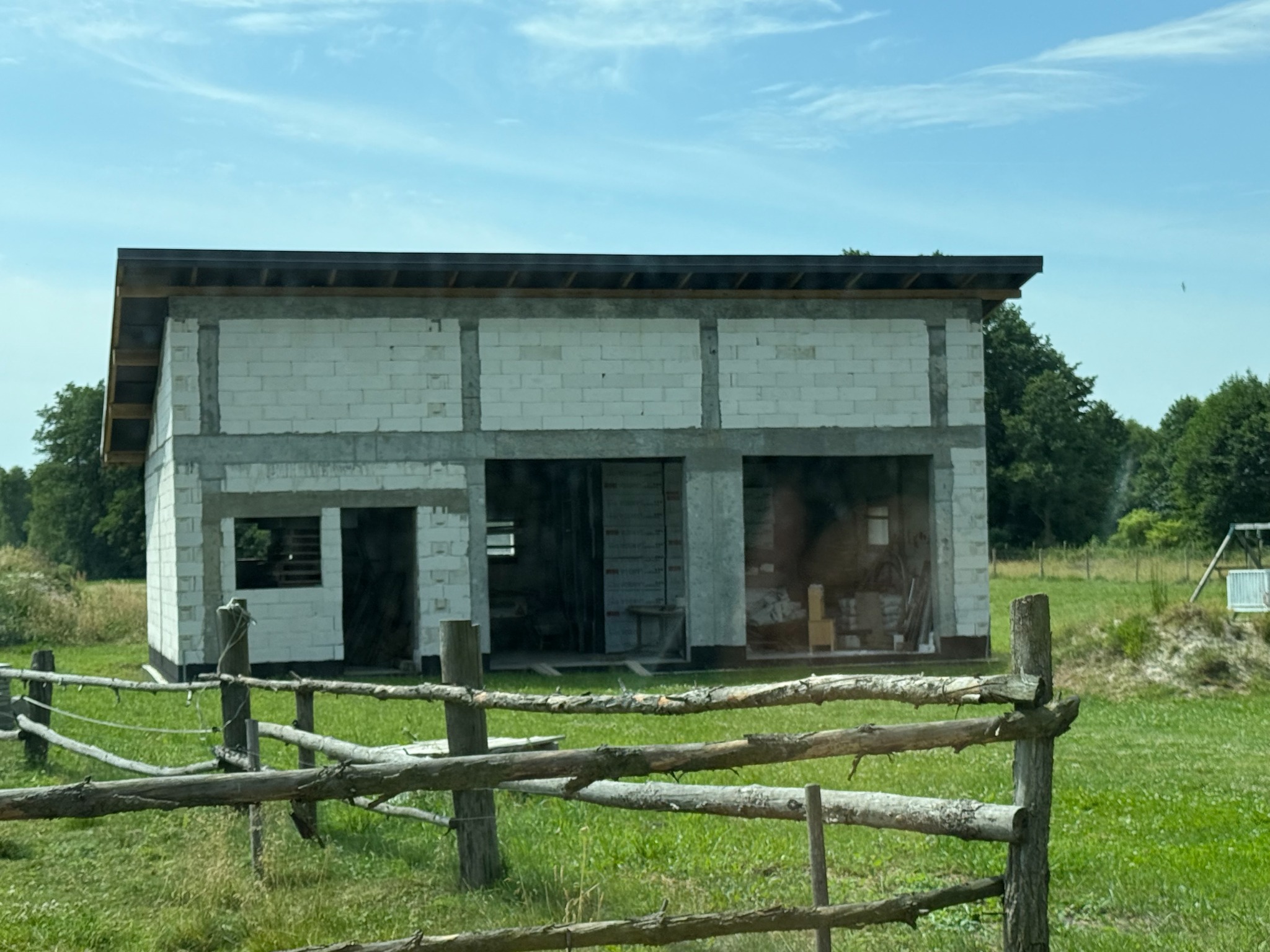 Niedokończona, parterowa hala z białych bloczków, z widoczną konstrukcją dachu, otoczona zielenią i drewnianym płotem. Budowa w toku, widoczne niedociągnięcia.