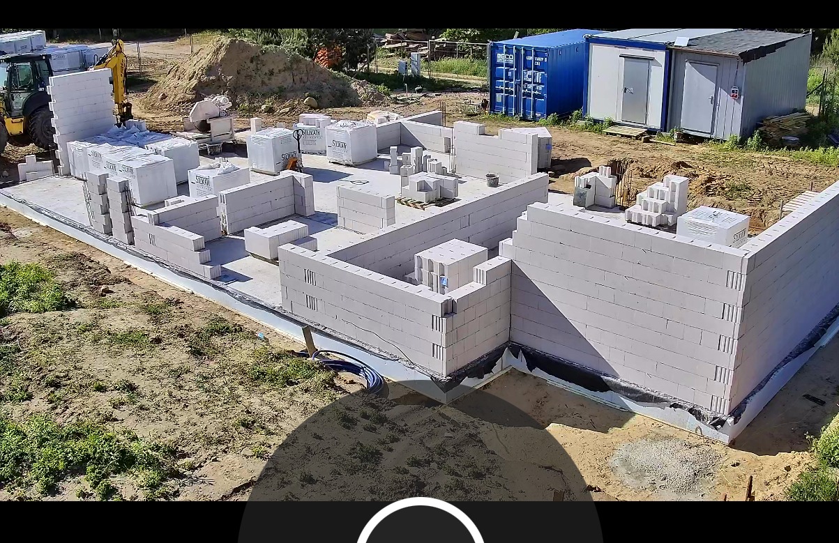 Fundamenty hali z bloczków silikatowych, częściowo wymurowane ściany, palety z materiałem budowlanym, teren budowy z kontenerami w tle.