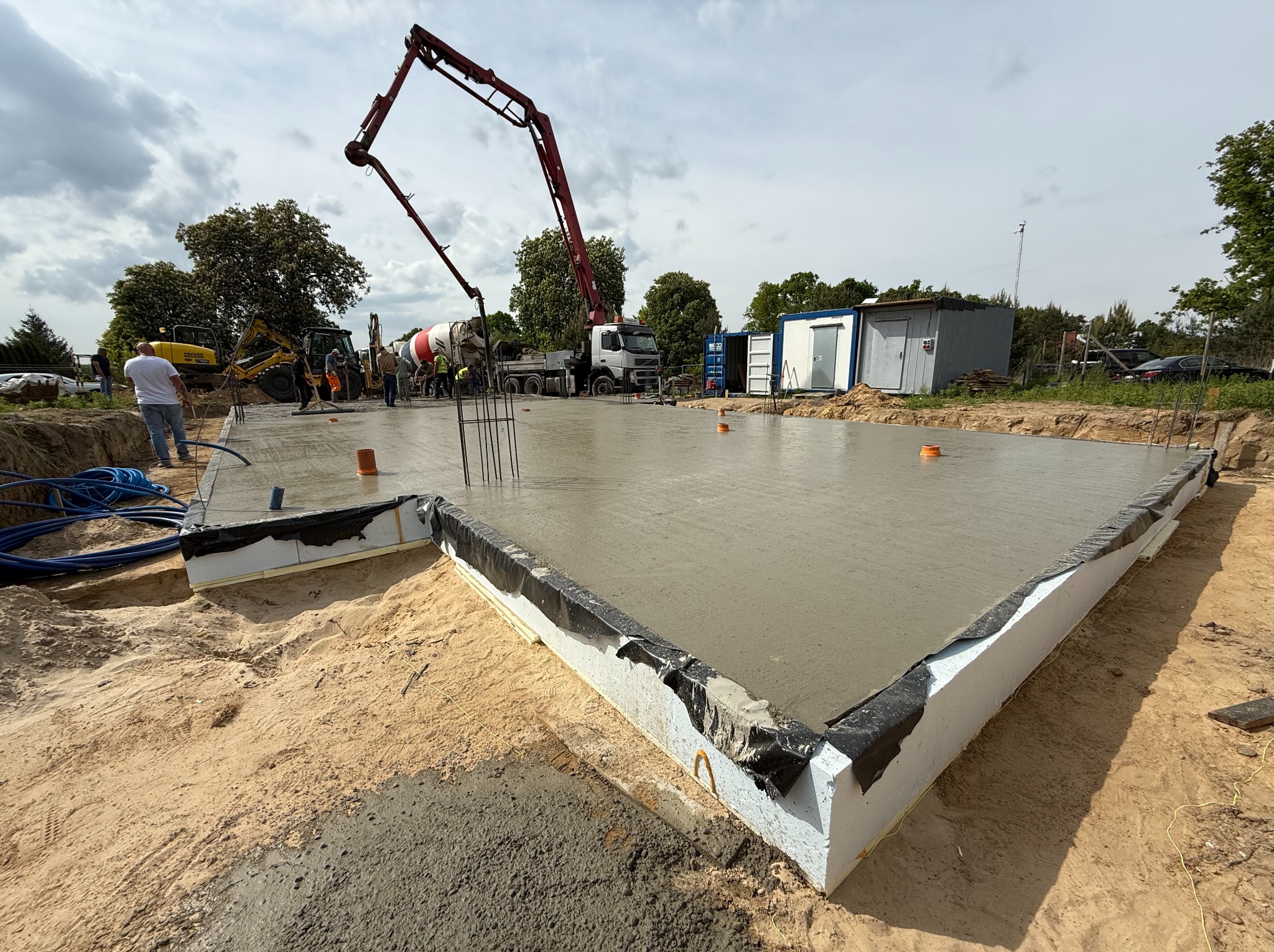 Wylany fundament pod halę z widoczną pompą do betonu, szalunkiem z izolacją i wystającymi prętami zbrojeniowymi. W tle betoniarka i ekipa budowlana.