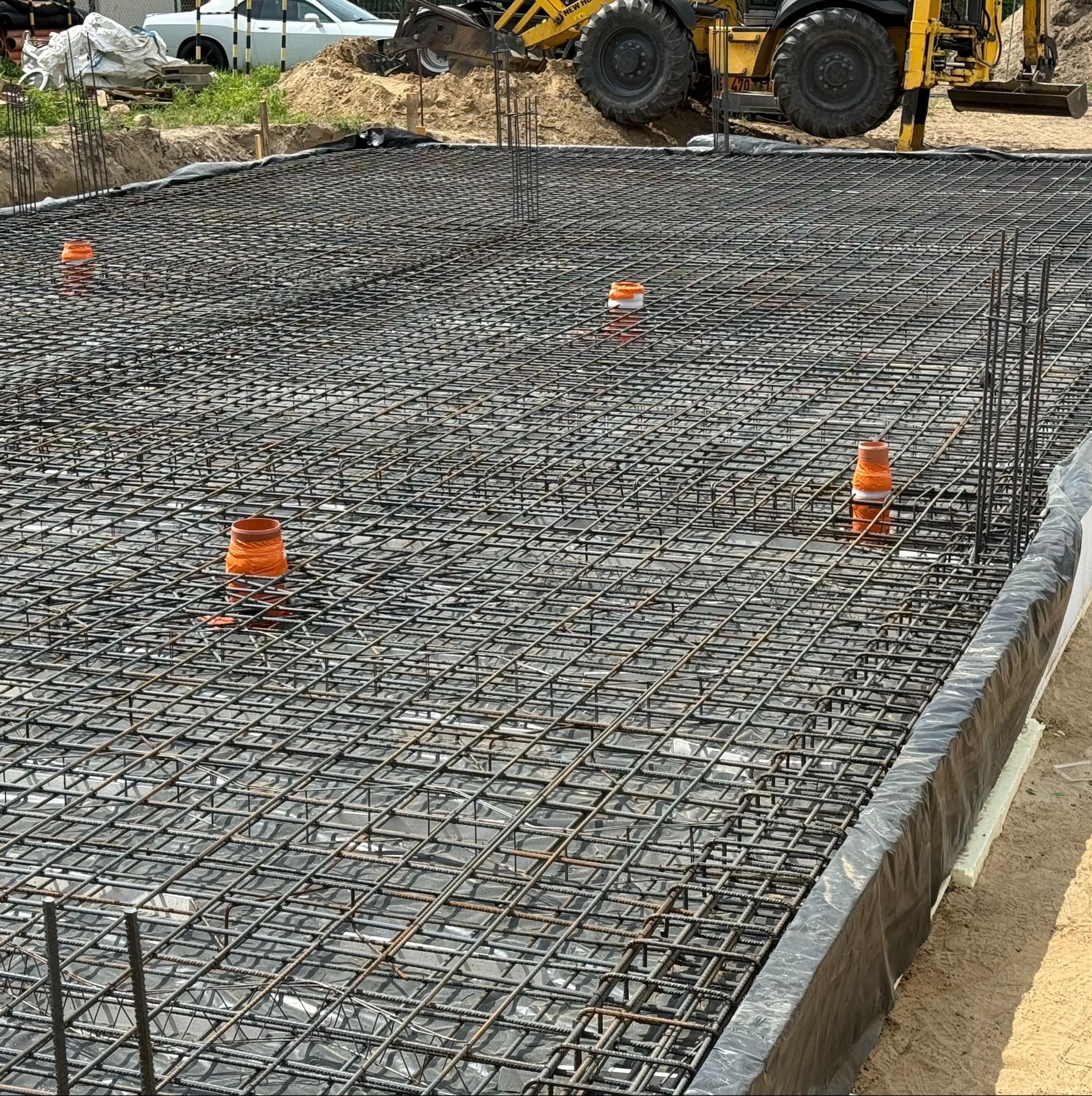 Zbrojenie fundamentu hali z widocznymi pionowymi prętami i pomarańczowymi rurami w tle z żółtą koparką. Siatka zbrojeniowa ułożona na folii izolacyjnej.