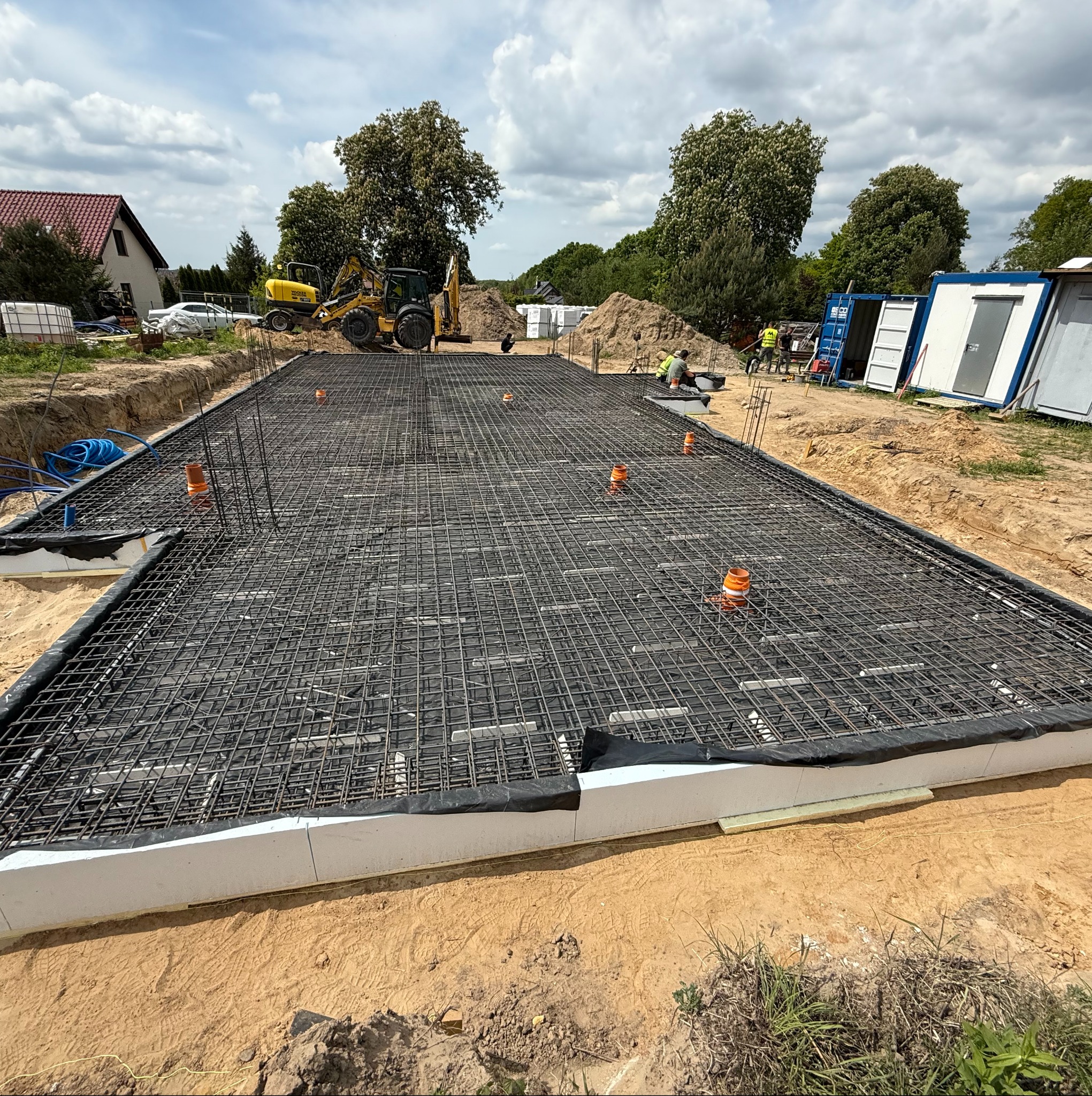 Zbrojenie fundamentu hali z widocznymi rurami instalacyjnymi, przygotowane do zalania betonem. W tle koparki i kontenery budowlane na placu budowy.