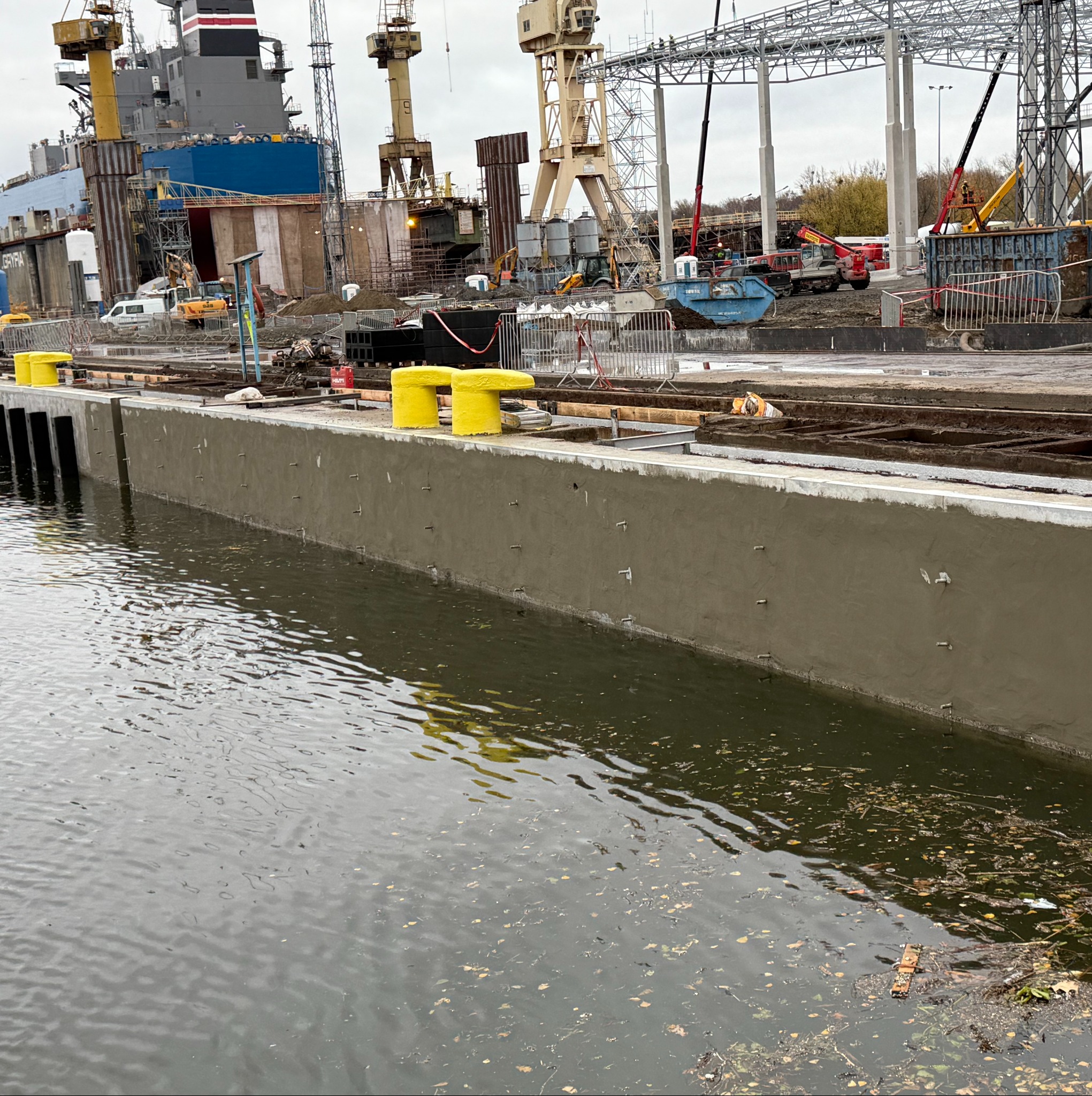 Betonowy nabrzeżny mur z żółtymi odbojami w Szczecinie, w tle budowa hali z dźwigami i elementami konstrukcji stalowej. Widok na wodę i plac budowy.
