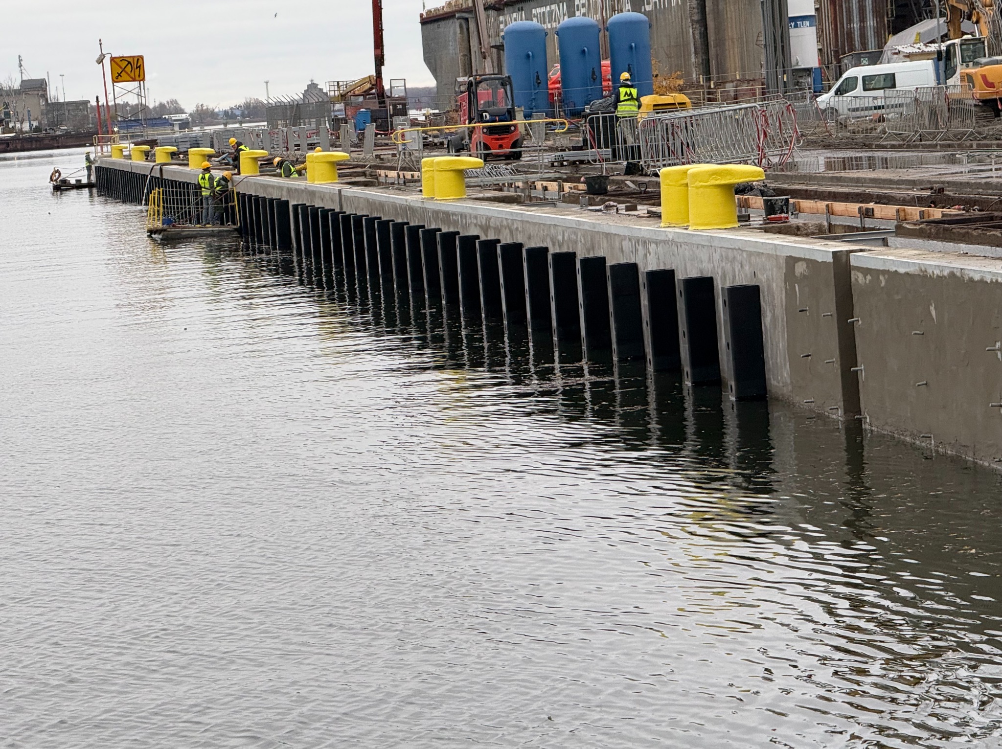 Nabrzeże portowe w trakcie budowy z żółtymi odbojami i pracownikami w kamizelkach, widok z poziomu wody. W tle widać sprzęt budowlany i elementy infrastruktury portowej.