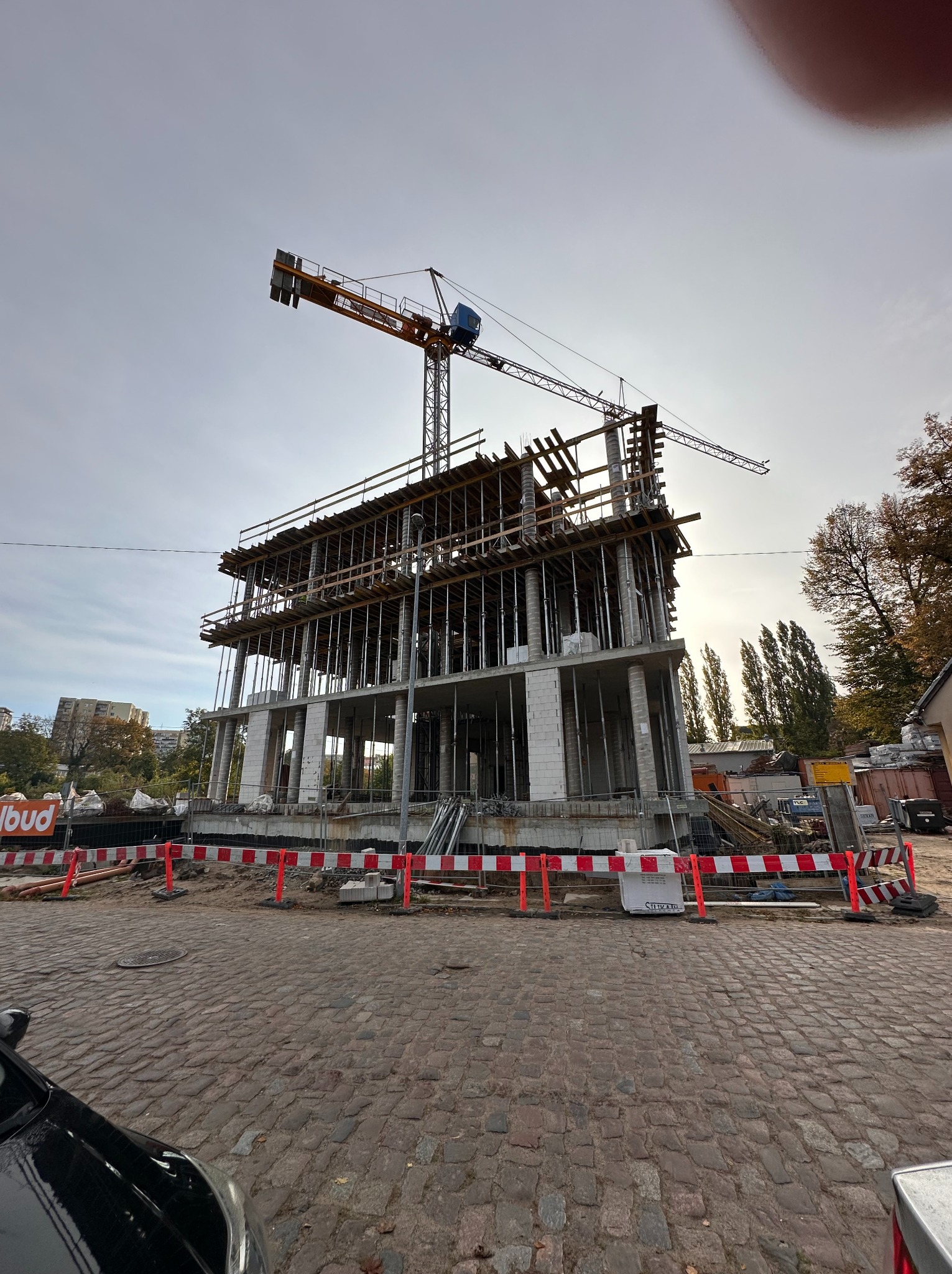 Wysoka konstrukcja w trakcie budowy z żurawiem na szczycie, widoczna z poziomu brukowanej ulicy, zabezpieczona barierkami. Częściowo widoczne drzewa w tle.