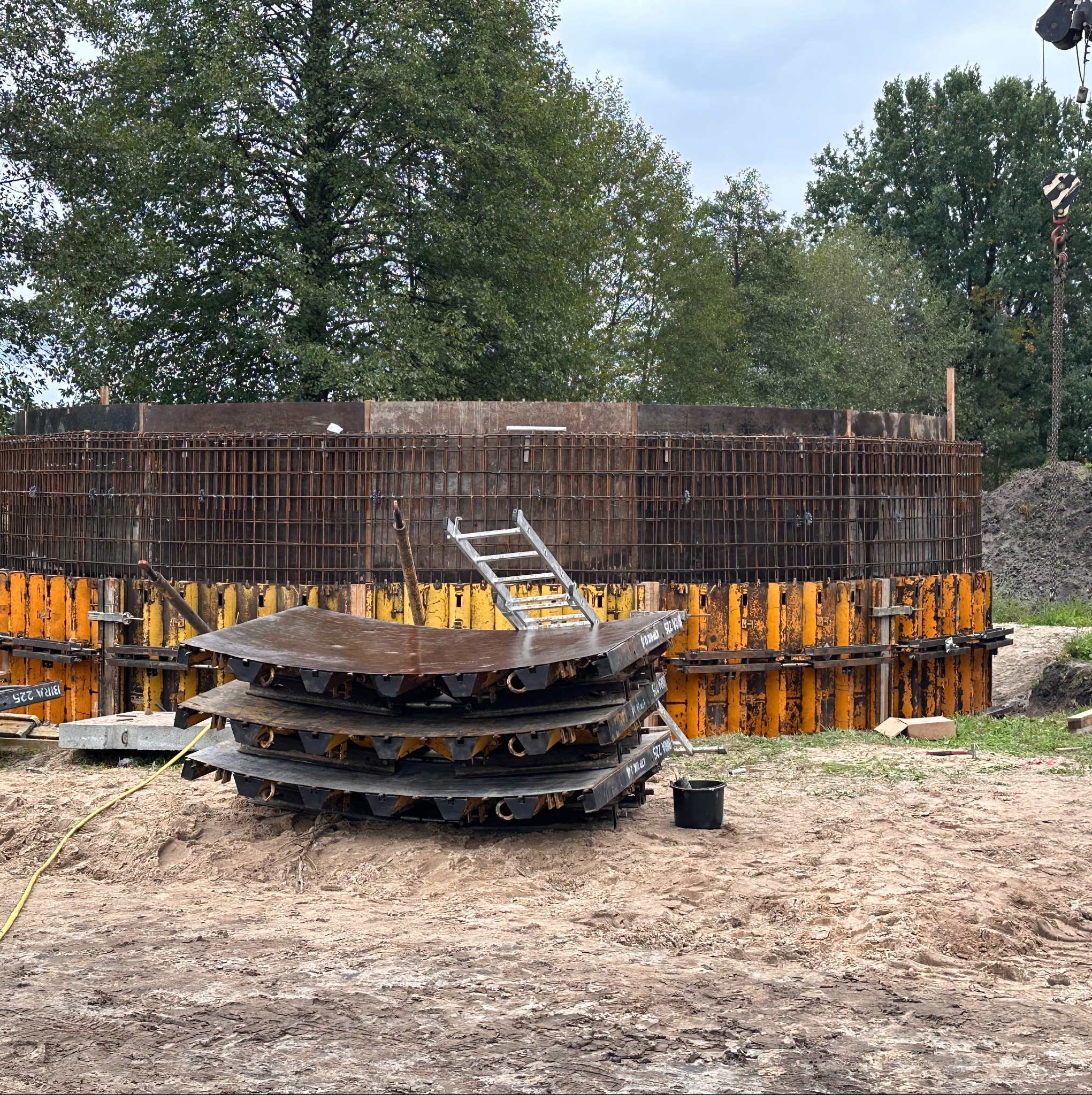 Okrągły szalunek zbrojeniowy w trakcie budowy, widoczne elementy konstrukcyjne i drabina. W tle drzewa i fragment żurawia.