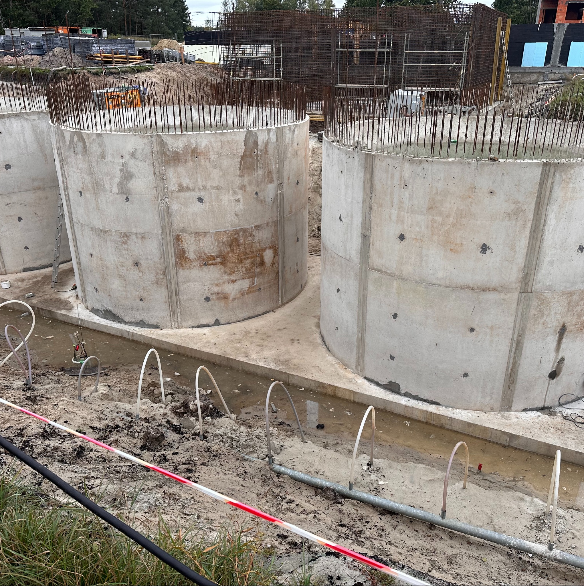 Budowa zbiorników betonowych z wystającymi prętami zbrojeniowymi, widoczne rury w ziemi, teren budowy z taśmami ostrzegawczymi. Surowy beton, etap budowy.