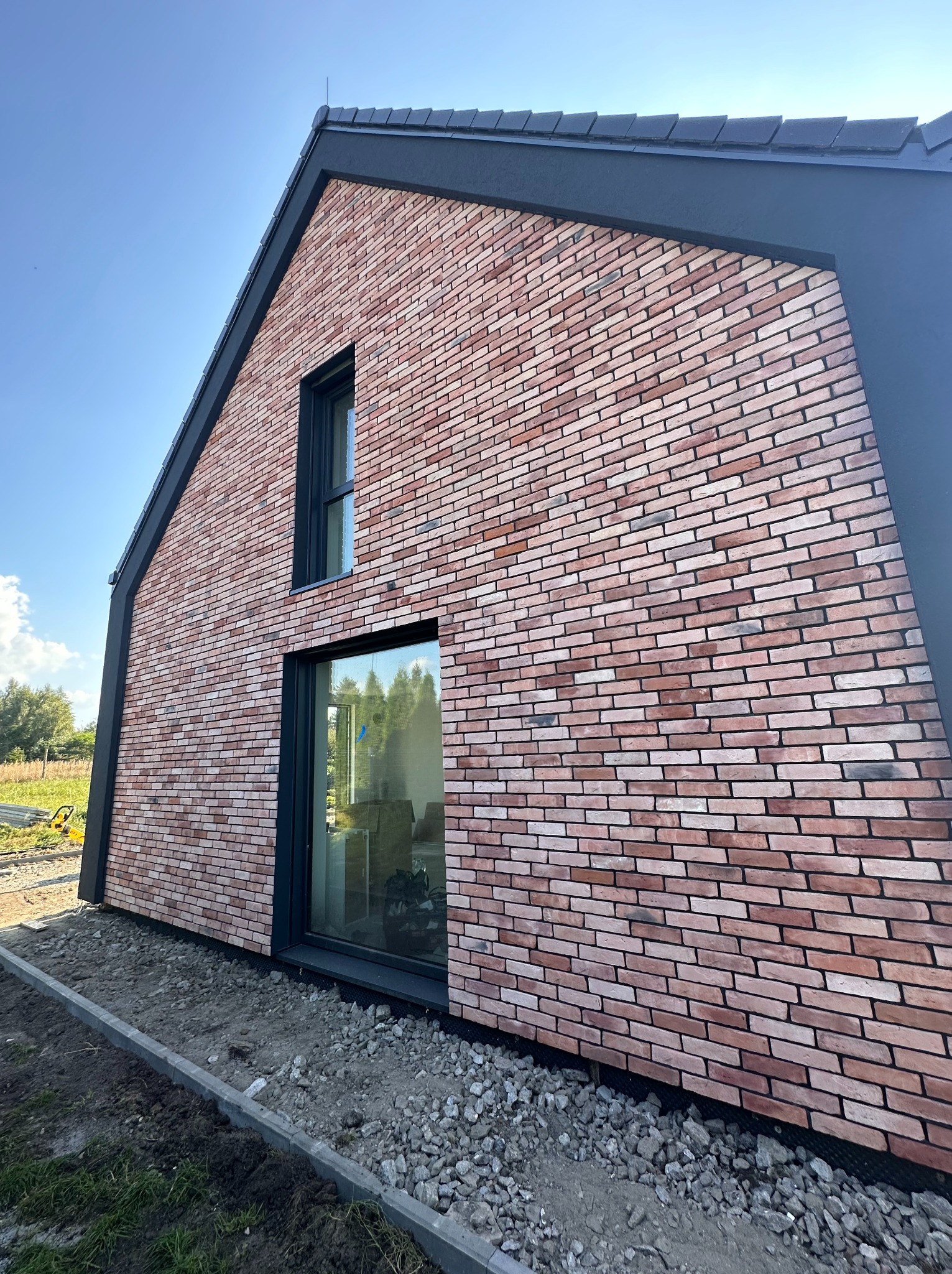 Nowoczesna hala z ceglaną elewacją i czarną stolarką okienną, widok z zewnątrz, w tle błękitne niebo i zieleń. Nietypowy kształt dachu.