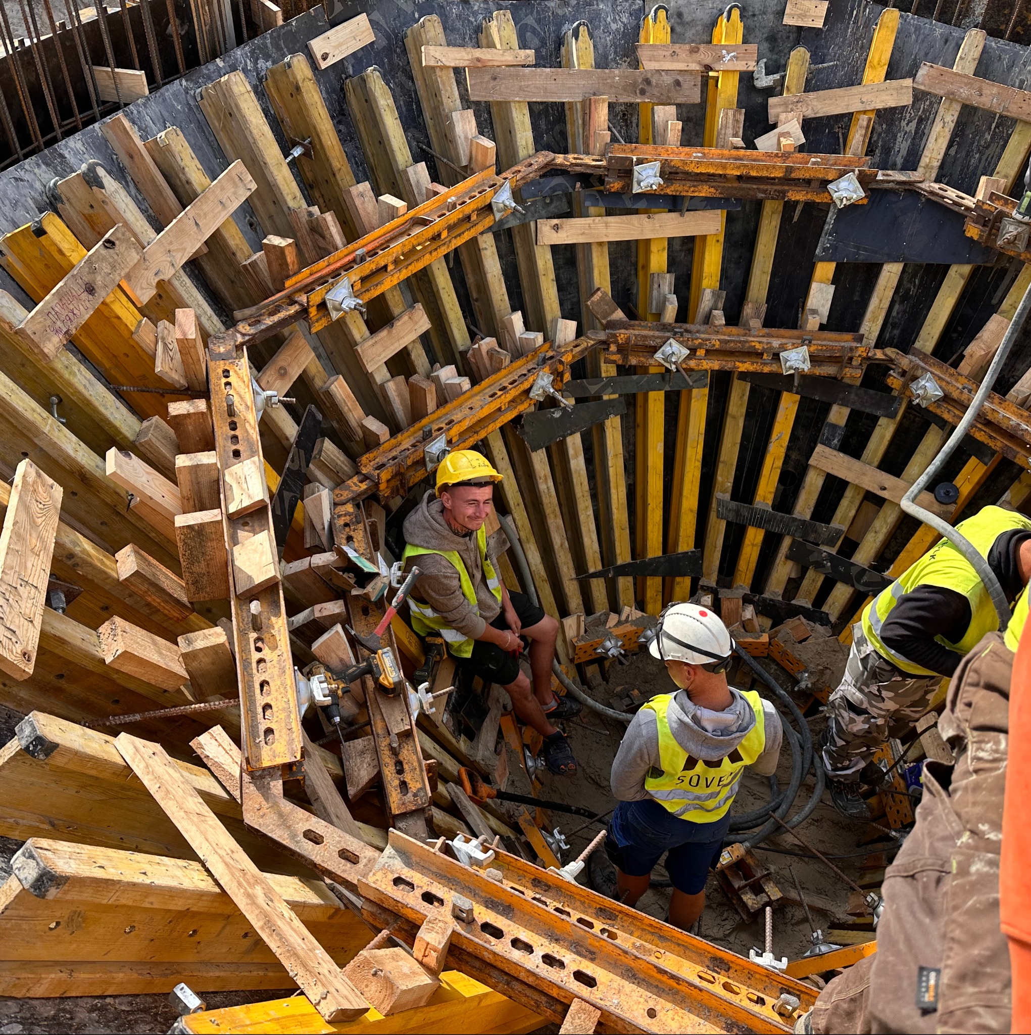 Budowa fundamentów: pracownicy w kasku i kamizelkach odblaskowych w wykopie z drewnianym szalunkiem, widok z góry na plac budowy.