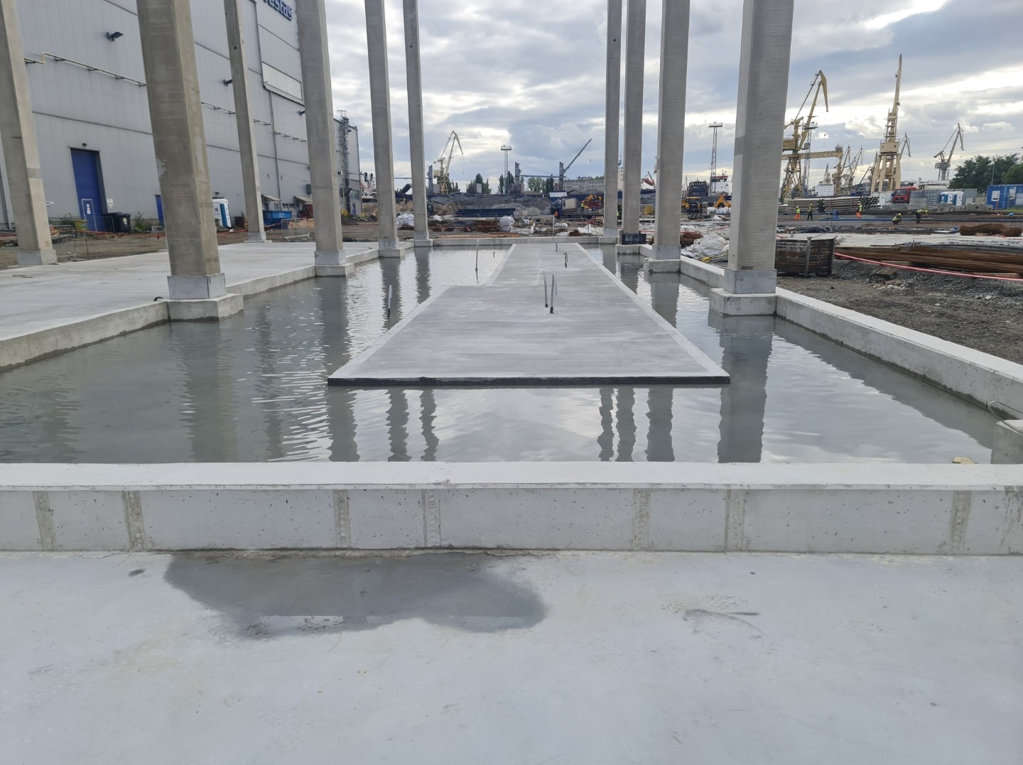 Wnętrze hali w budowie z betonowymi kolumnami i płytą fundamentową częściowo zalaną wodą. W tle widoczne dźwigi i elementy konstrukcyjne.