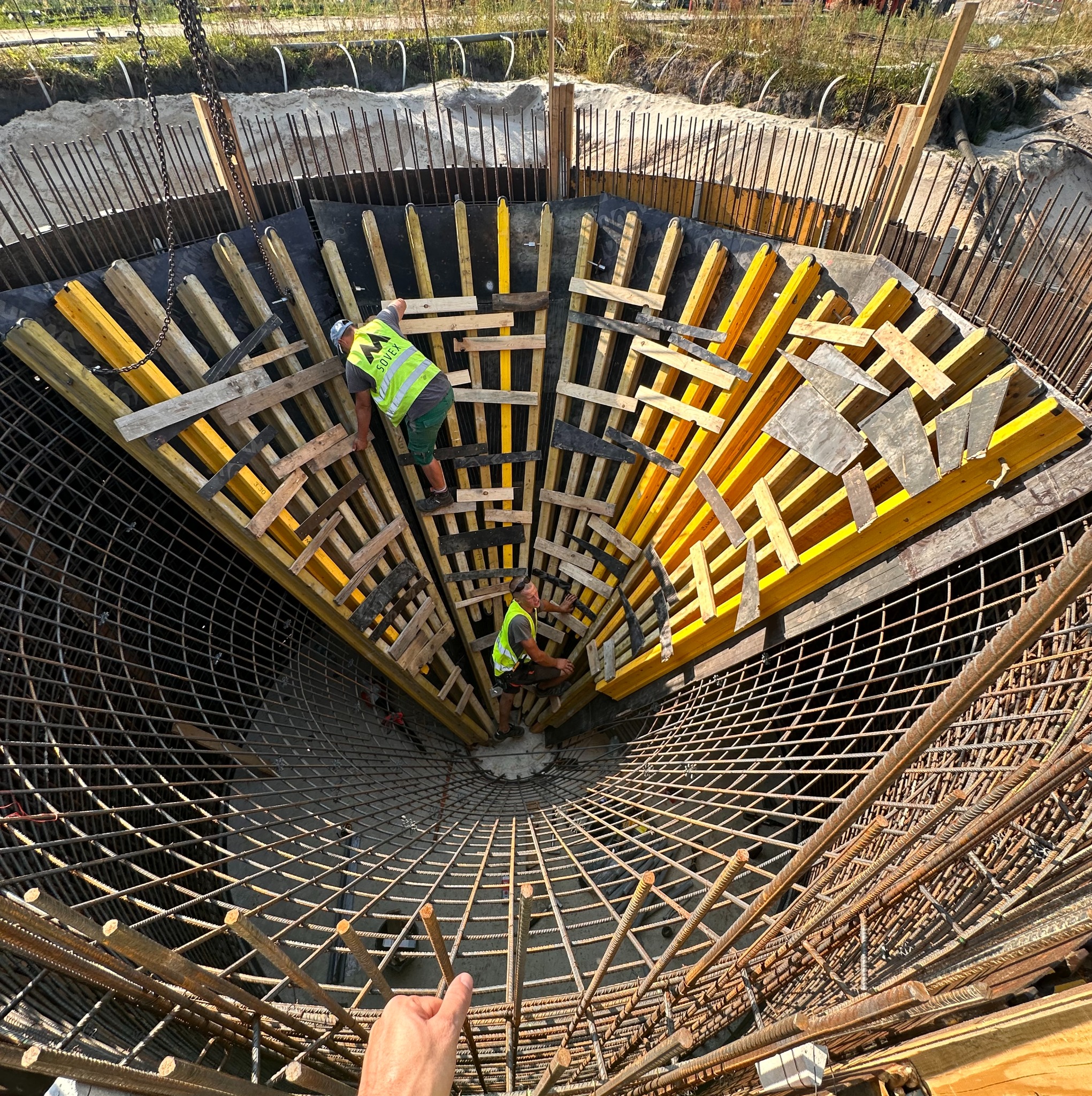 Dwóch pracowników w żółtych kamizelkach montuje szalunek i zbrojenie w okrągłym wykopie pod fundament. Widok z góry, z ręką w kadrze.