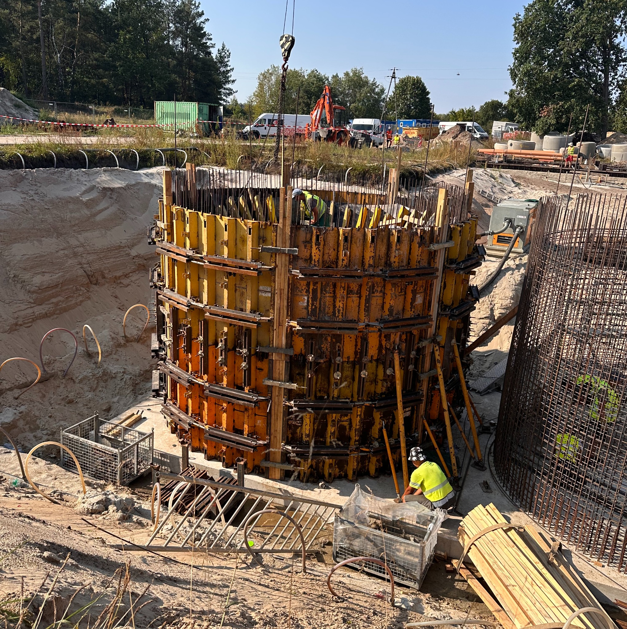 Budowa cylindrycznej konstrukcji z żółtymi szalunkami i stalowym zbrojeniem na placu budowy. W tle widoczny sprzęt budowlany i pracownicy w odblaskowych kamizelkach.