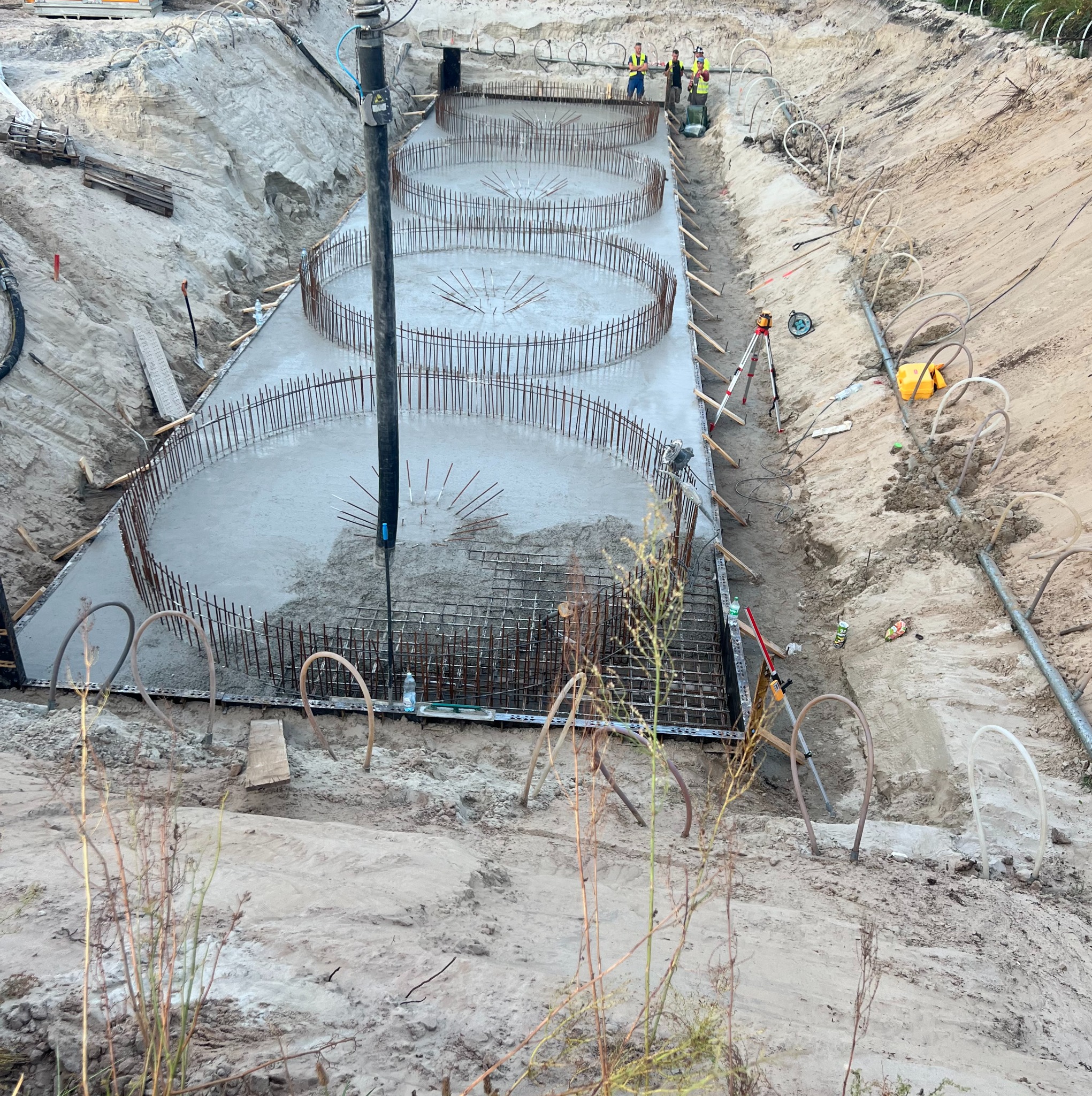 Wykop z trzema okrągłymi fundamentami zbrojonymi stalą, zalewanymi betonem. W tle pracownicy budowlani i sprzęt pomiarowy. Widok z góry na plac budowy.