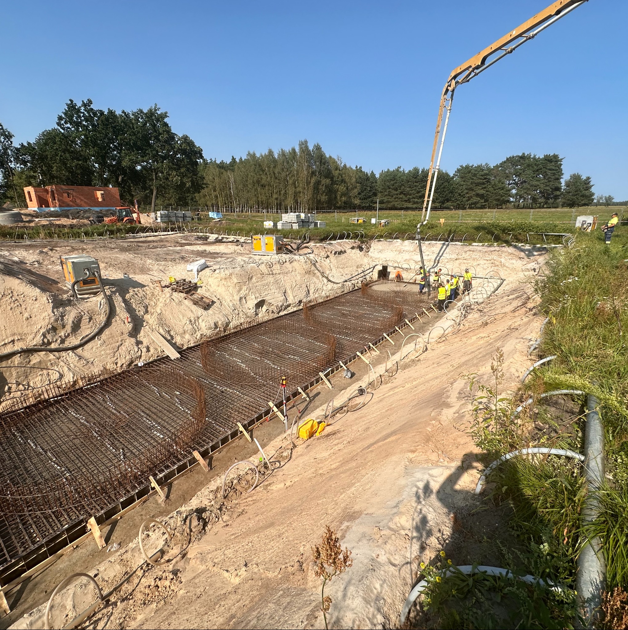 Zbrojenie fundamentów hali w wykopie, widok z góry. Na budowie obecni pracownicy w żółtych kamizelkach, pompa do betonu. W tle drzewa i częściowo wybudowany budynek.